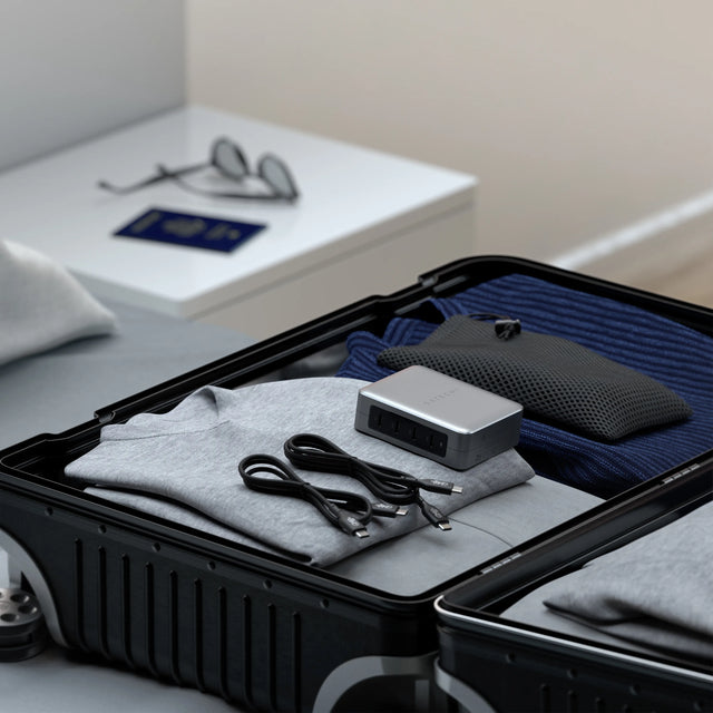 Satechi 145W travel charger in space gray and two USB-C cables packed in an open suitcase with folded clothes, a passport and sunglasses on a nightstand nearby.
