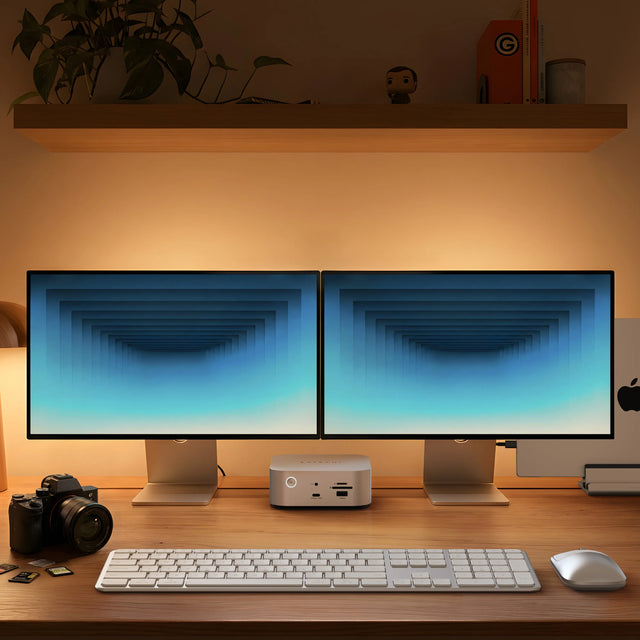 Satechi Thunderbolt 5 CubeDock™ with SSD Enclosure sitting between two monitors on a wooden desk, with a white keyboard, mouse, camera and SD cards in front and a laptop docked at the right.