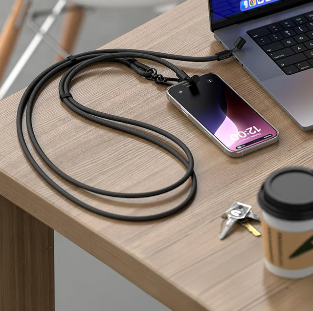 Black braided Satechi OntheGo™ USB-C Lanyard Cable attached to an iPhone and plugged into a laptop on a wooden desk, next to keys and a coffee cup.