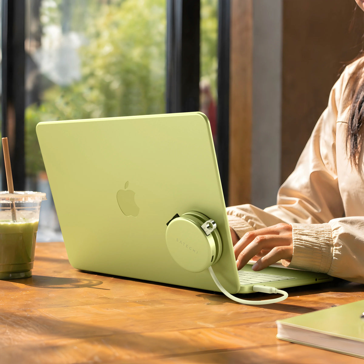 A Citrus OntheGo™ 5-in-1 Multiport Adapter attached to Citrus MacBook Neo, shown on a wooden table in a cafe setting with a person typing on the laptop.