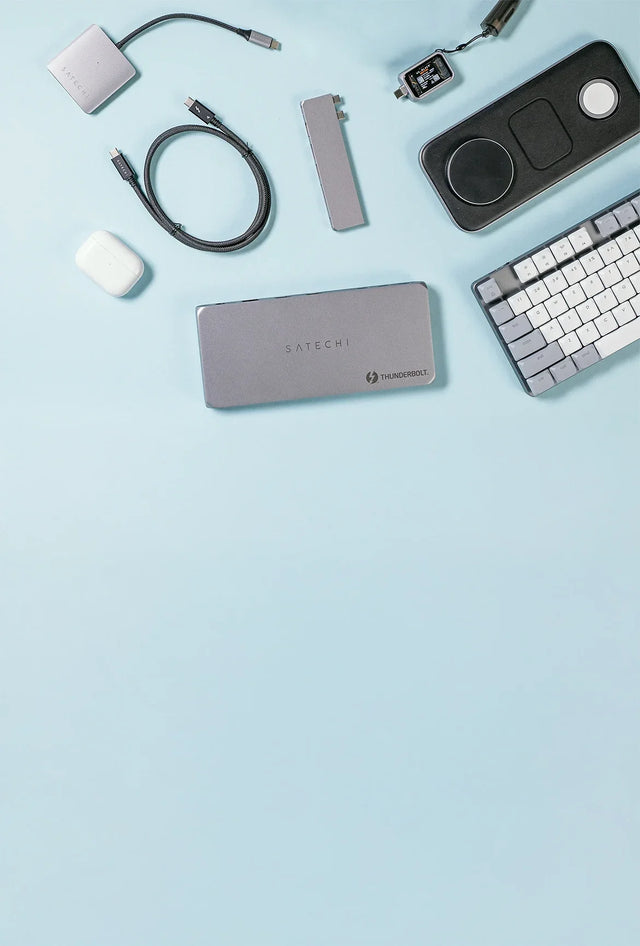 Overhead view of Satechi accessories on a light blue surface: a Thunderbolt dock, Pro Hub Slim, braided Thunderbolt 4 cable, compact hub, Qi2 Trio Wireless Charging Pad, SM3 Slim Mechanical Backlit Bluetooth Keyboard, and AirPods case.