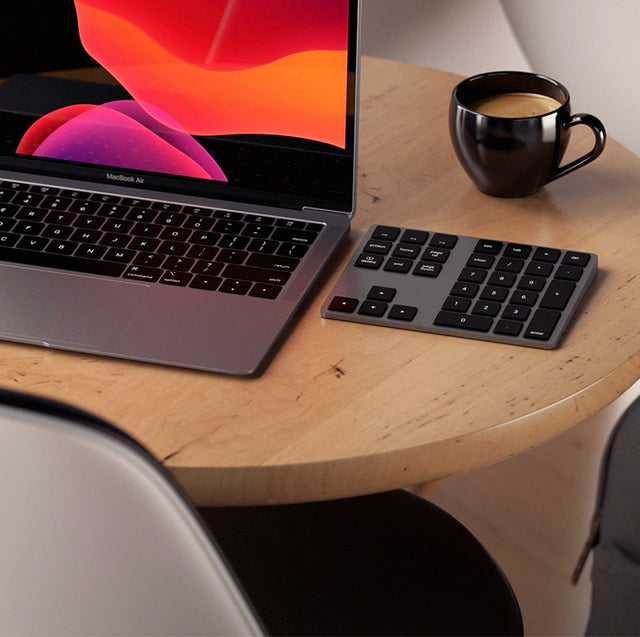 Satechi Bluetooth Extended Keypad in space gray next to a MacBook Air and a cup of coffee on a round wooden table.