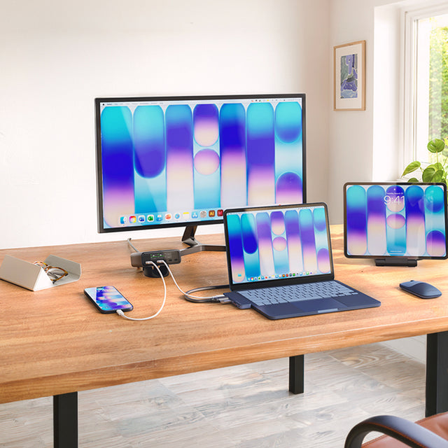 Satechi USB-C Snap Hub in Indigo connected to an Indigo MacBook Neo, with the OntheGo Foldable Stand and Hub holding an iPad and an Indigo EX Mouse. A ChargeView 240W Charger charges an iPhone and MacBook Neo, with a Findall Glasses Case nearby on the desk.