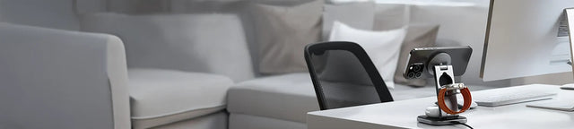 Satechi 3-in-1 Foldable Qi2 Wireless Charging Stand on a white desk charging an iPhone and Apple Watch, in a home office with a gray sofa in the background.