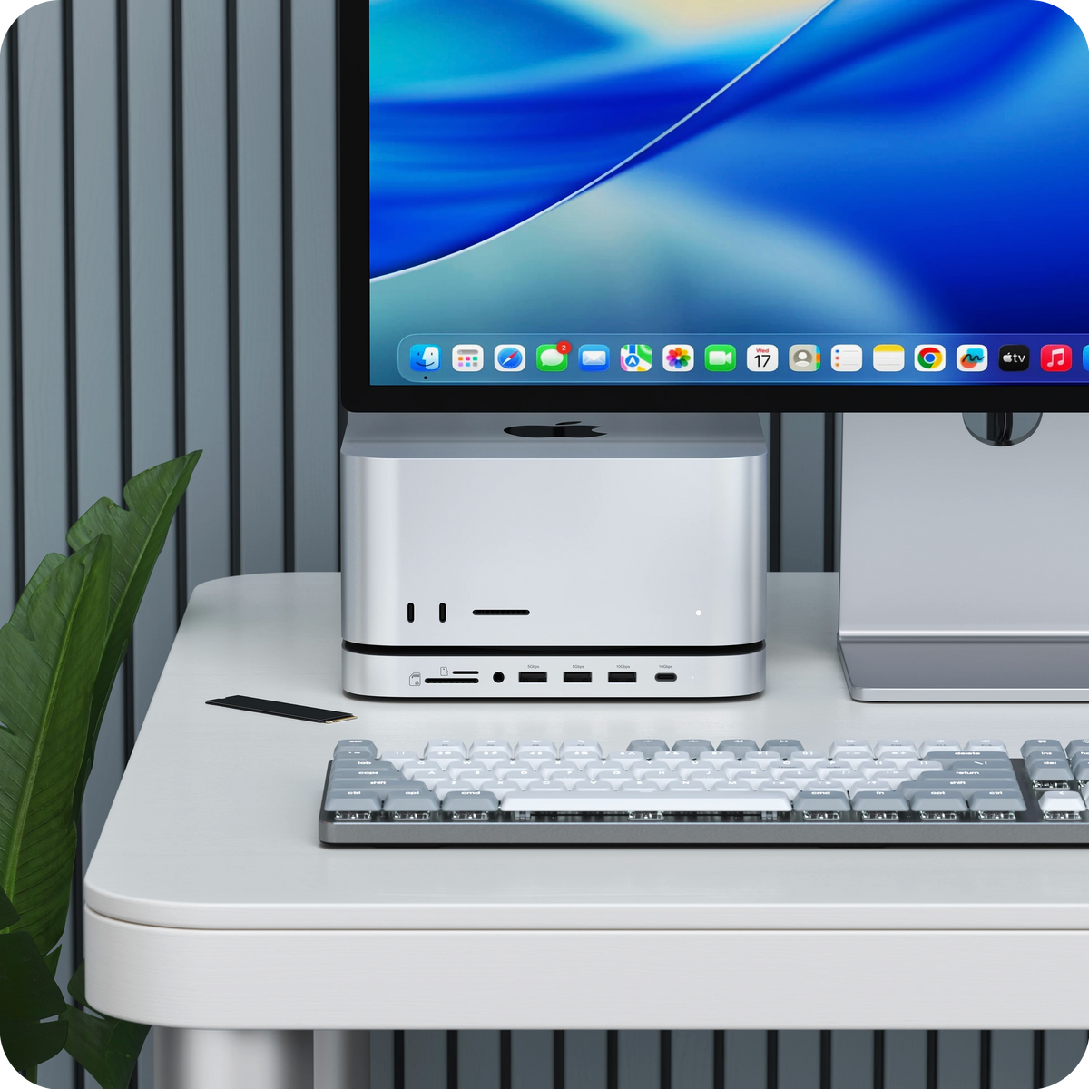 Stand & Hub for Mac Studio M5 with NVMe SSD Enclosure under a Mac Studio on a desk, with a monitor and keyboard