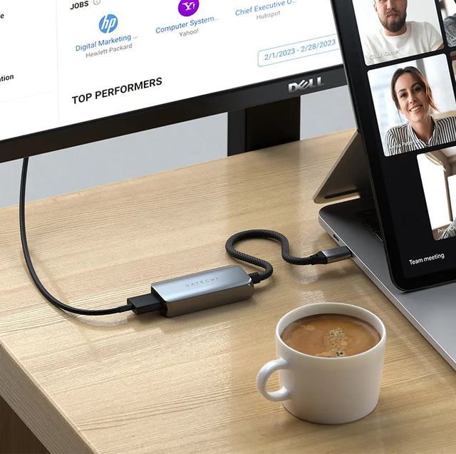 Satechi USB-C to HDMI 2.1 8K Adapter connecting a laptop to a Dell monitor on a wooden desk, beside a cup of coffee and a tablet showing a video call.