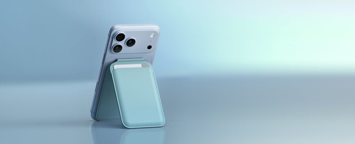 A  iPhone 18 Pro in Glacier propped up in portrait on a glossy light-blue surface, held at an angle by the Satechi Vegan-Leather Magnetic Wallet Stand in Light Blue folded out into stand mode behind it. The stitched wallet pocket holds a single white card, its top edge visible above the opening. A magsafe wallet stand that doubles as a kickstand.