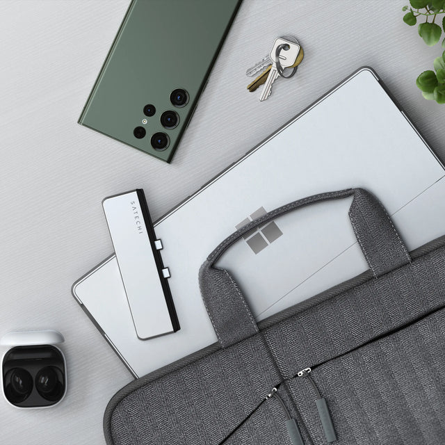 Satechi Dual USB-C Hub for Surface Pro 9 in silver on a Surface Pro, laid on a desk with a laptop bag, keys, an Android phone and wireless earbuds.