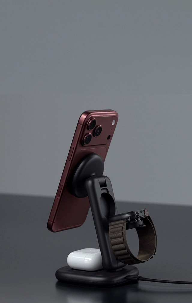 Satechi 3-in-1 Foldable Wireless Charging Stand with Qi2 25W in black, charging an iPhone 18 Pro Max in Burgundy on its magnetic stand, an Apple Watch with a dark green loop band on the watch charger, and AirPods in their case on the base. The Qi2 charging stand sits on a dark gray surface with its power cable running off to the side.