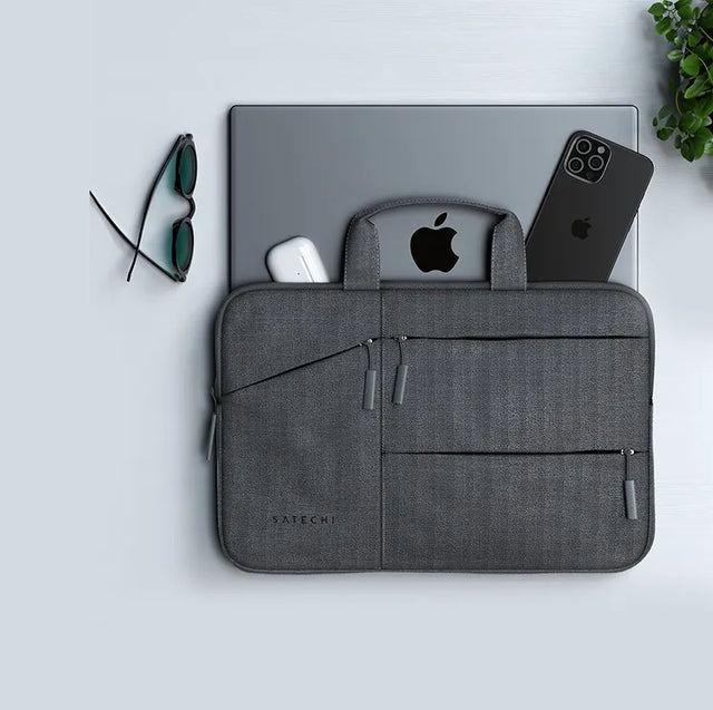 Gray Satechi laptop carrying case with handles and zip pockets, with a MacBook, iPhone and AirPods case tucked behind it, next to sunglasses and a plant.