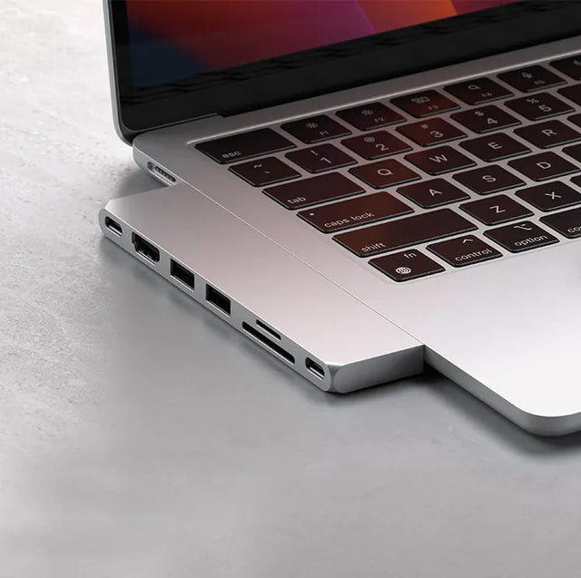 Silver Satechi USB-C multiport hub attached to the side of a MacBook Pro, showing USB-C, HDMI, two USB-A, SD and microSD card slots, and a second USB-C port.