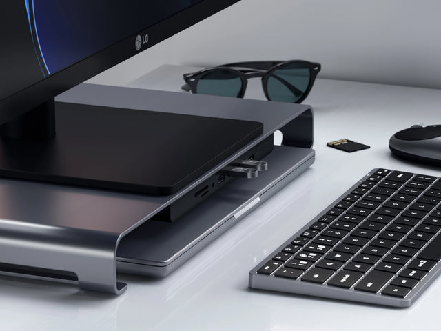 Satechi USB-C Monitor Stand Hub XL in space gray under an LG monitor, with USB flash drives in its front ports, next to a wireless keyboard, mouse and SD card.