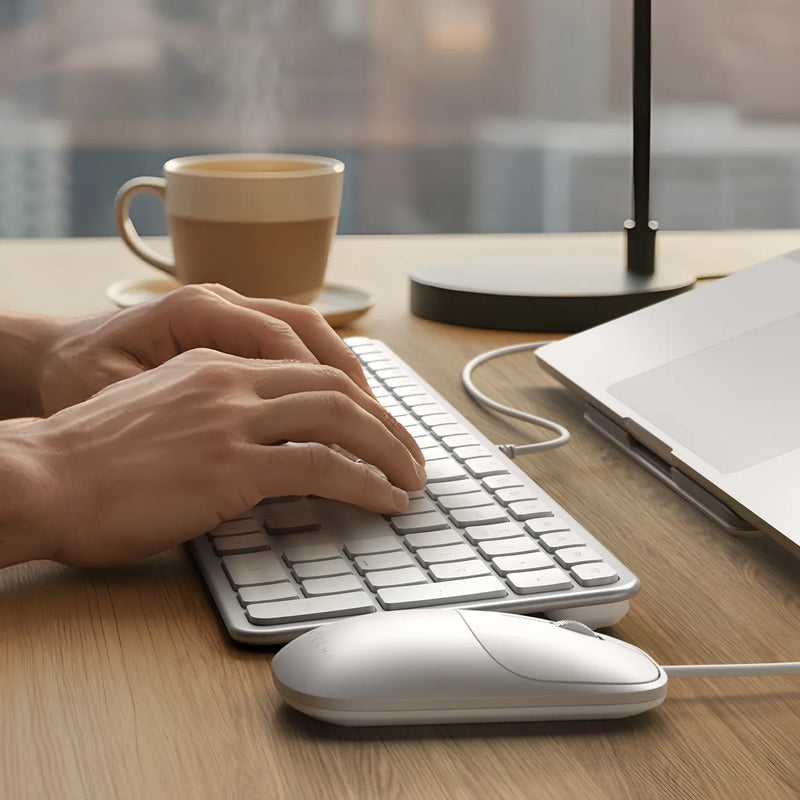 Hands typing on a Satechi Slim CLX3 wired keyboard in white with the matching wired mouse, next to a laptop and a cup of coffee.