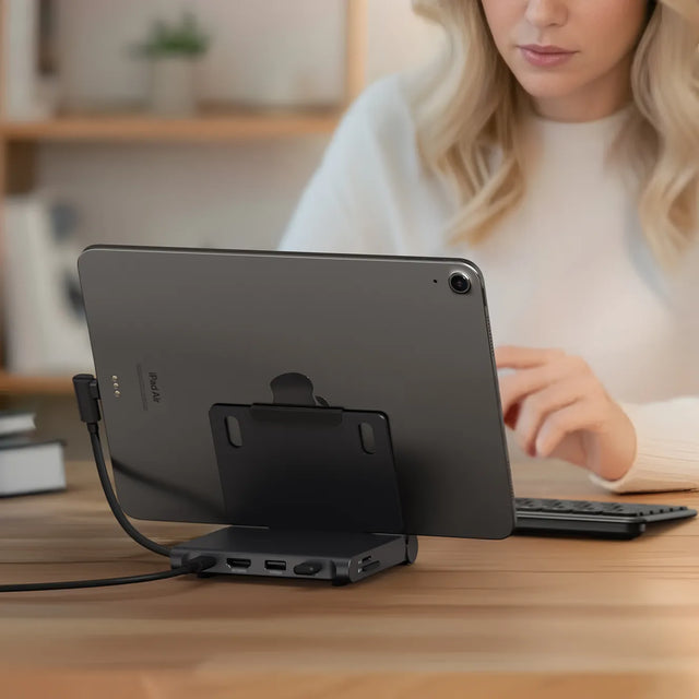 iPad connected to the Satechi OntheGo™ Foldable Stand Hub on a desk, with the hub’s USB‑C and data ports visible while a person types on a keyboard in the background.