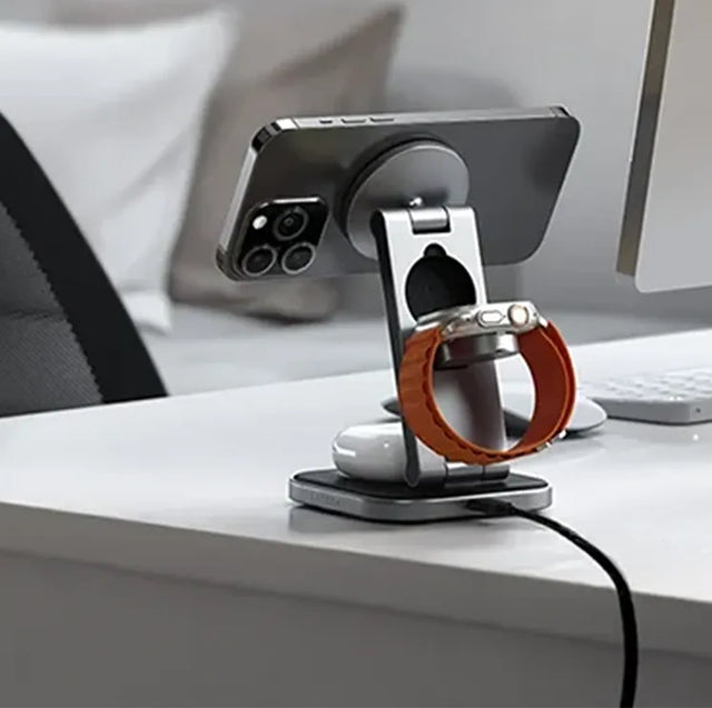 Satechi 3-in-1 magnetic wireless charging stand on a desk, charging an iPhone, an Apple Watch with an orange band and AirPods.
