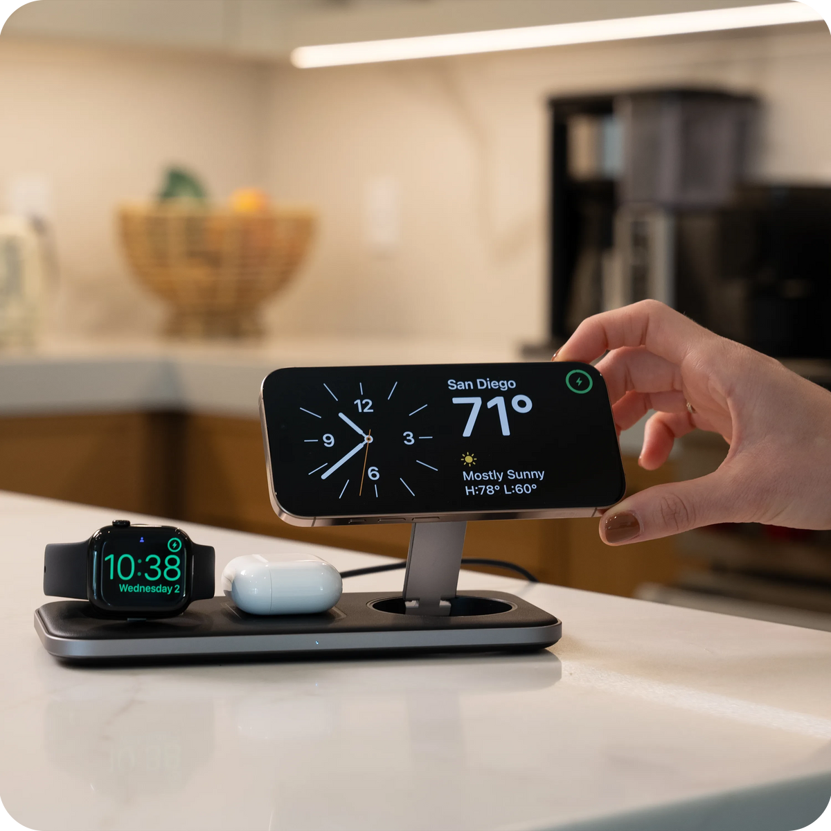 Hand placing an iPhone on the Qi2 Trio Wireless Charging Pad on a kitchen counter, with Apple Watch and AirPods charging