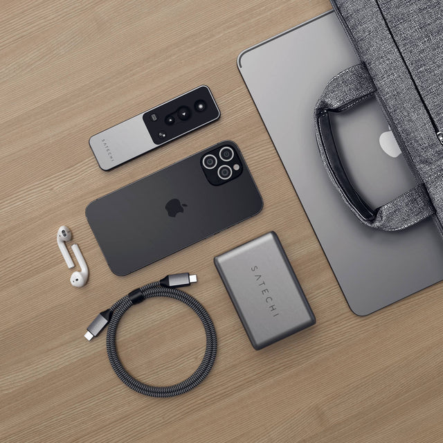 Overhead view of the Satechi R1 Bluetooth Presentation Remote on a wooden desk with an iPhone, AirPods, a Satechi USB-C charger, a braided USB-C cable and a laptop in a sleeve.