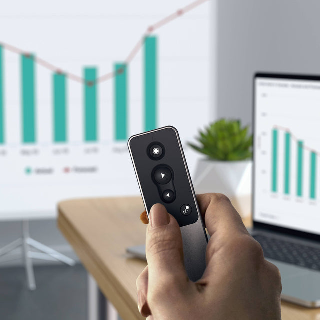 Hand holding the Satechi R1 Bluetooth Presentation Remote in front of a projected bar chart, with a laptop showing a matching chart on a desk.