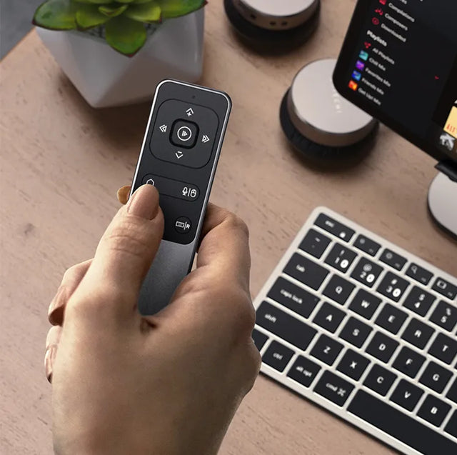 Hand holding the Satechi R2 remote above a desk with a wireless keyboard, speakers and a tablet.