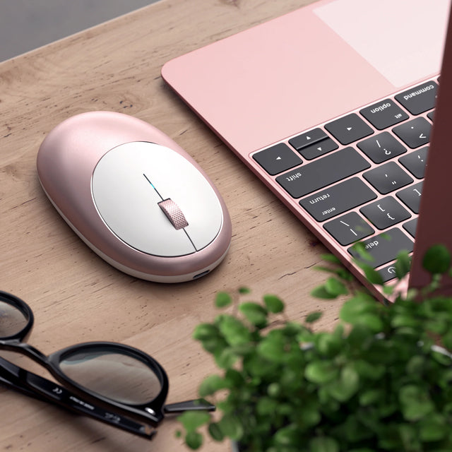 Satechi M1 Wireless Mouse in rose gold on a wooden desk beside a rose gold MacBook, glasses and a plant.