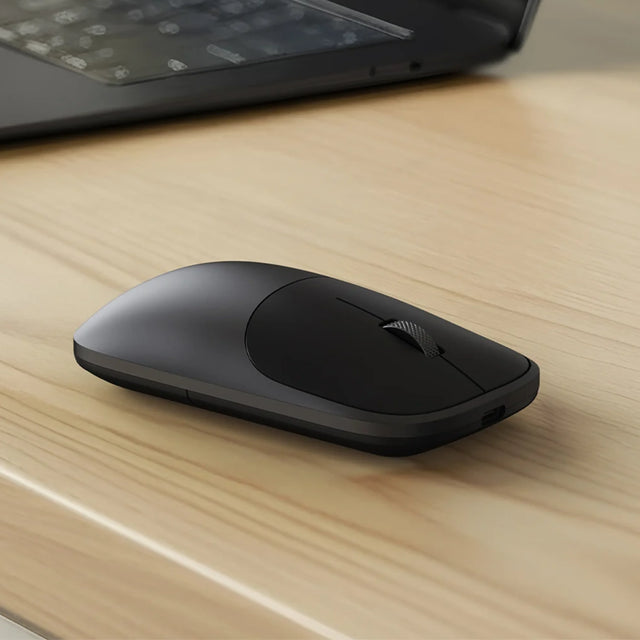 Black Satechi Slim EX Mouse on a wooden desk next to a laptop.
