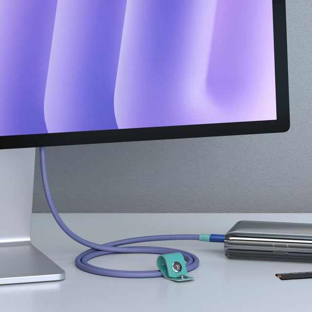 Purple Satechi right-angle USB-C to USB-C data and charge cable with a teal cable strap, connecting a monitor to a laptop on a desk.